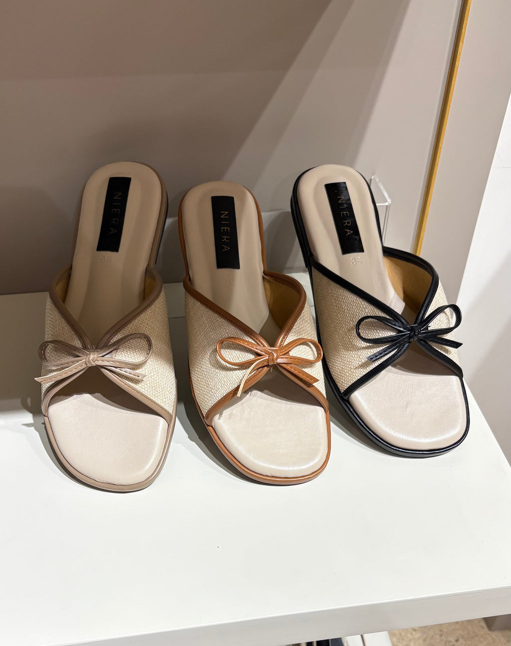Pair of beige mules with bow detail on a white shelf.