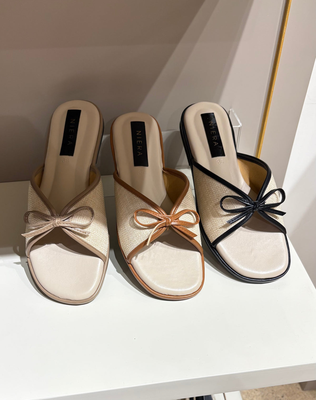 Pair of beige mules with bow detail on a white shelf.
